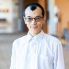 Smiling Asian man with glasses in white shirt; professional headshot, blurred office background